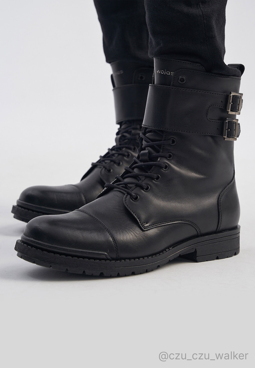 Czarne męskie bikery Wojas 24034-51 ze skóry licowej, z podwójnymi klamrami, sznurowane, za kostkę, na grubej podeszwie, prezentowane na nogach w ciemnych spodniach.