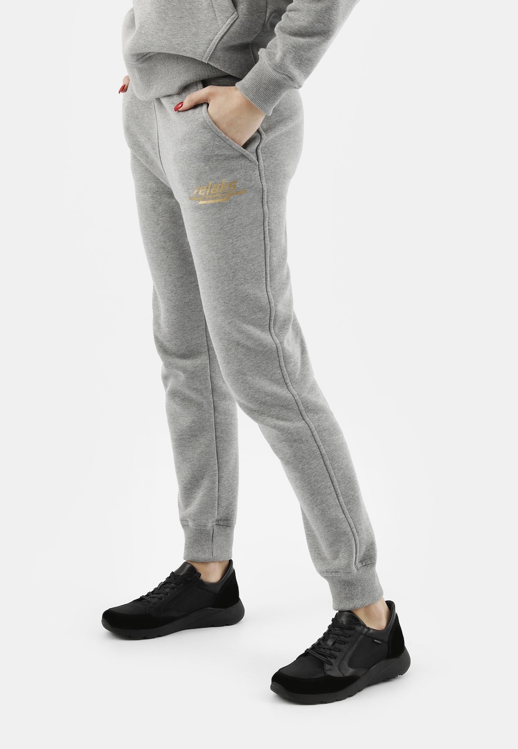 Popielate spodnie dresowe damskie model 98024-80 widoczne na nodze modelki z dłonią w kieszeni. Wykonane z bawełny, z małym logo. W zestawieniu z czarnymi butami Relaks.