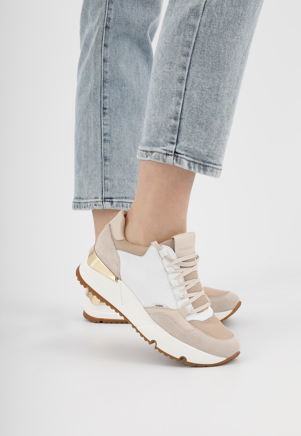 Damskie sneakersy Wojas 46137-89 w biało-beżowej kolorystyce ze sznurowaniem, noszone z jasnymi jeansami. Buty mają charakterystyczną podeszwę z brązowym spodem.