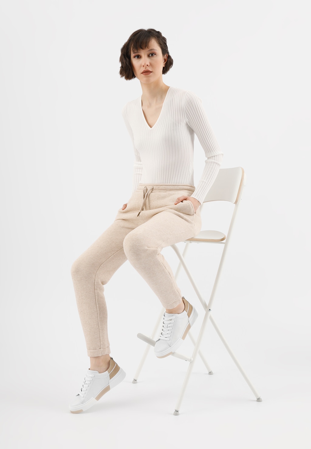 Kobieta w białym sweterku i beżowych spodniach siedzi na krześle, prezentując białe trampki damskie Wojas model 46121-54 z charakterystycznymi beżowymi akcentami.