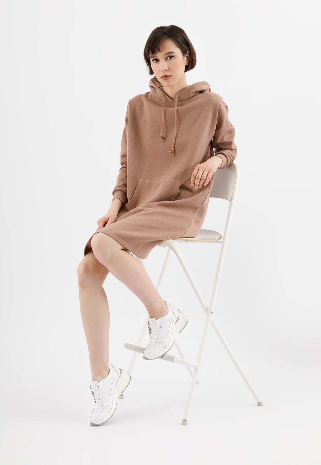 Modelka w beżowej sukience z kapturem siedząca na krześle, prezentująca białe sneakersy damskie Wojas model 46115-79 na podwyższonej podeszwie, widoczne na jej stopach.