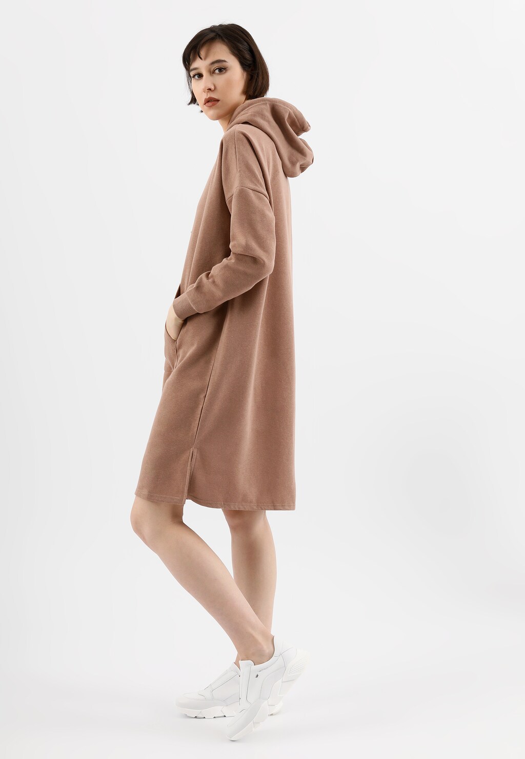 Modelka w beżowej, luźnej sukience z kapturem prezentująca białe sneakersy wsuwane Wojas 46119-59 na grubej podeszwie, w casualowej stylizacji.