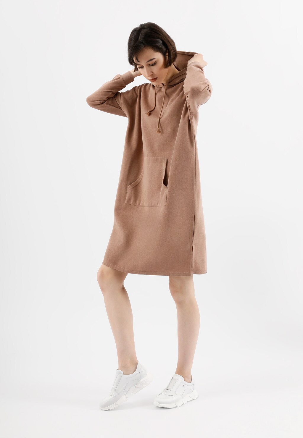 Modelka w beżowej sukience z kapturem prezentuje białe wsuwane sneakersy damskie Wojas model 46119-59 na grubej podeszwie. Sportowa, casualowa stylizacja na białym tle.