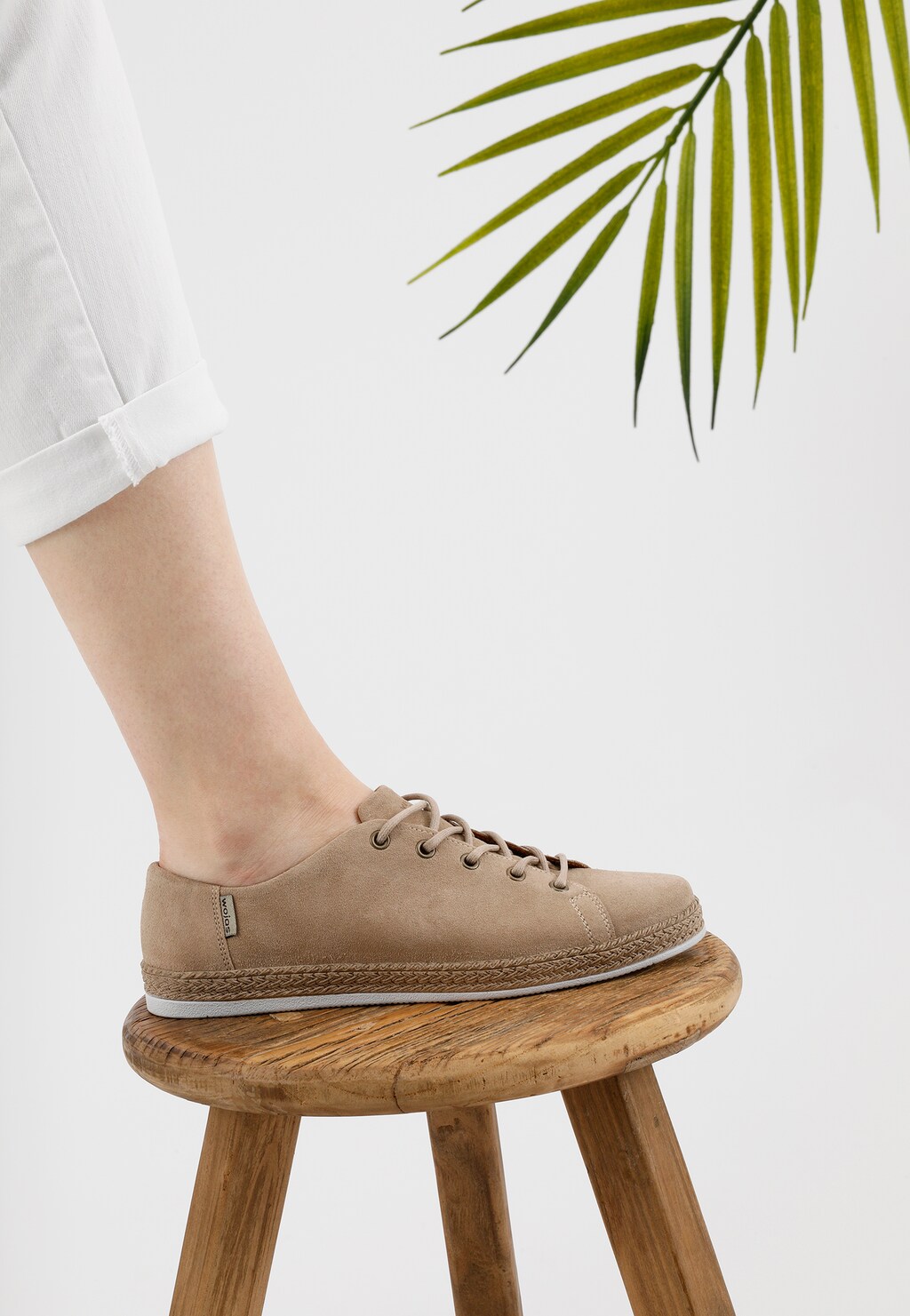 Beżowe półbuty damskie Wojas model 46150-64 wykonane z dwoiny, prezentowane na nodze modelki, z plecioną podeszwą i minimalistycznym designem, obok liścia palmy.