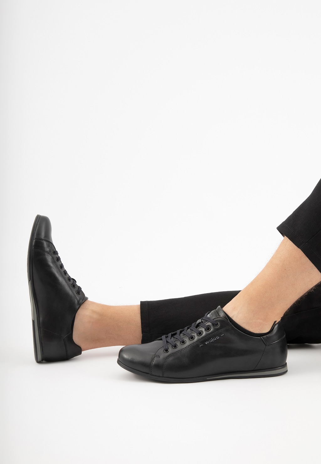 Czarne minimalistyczne sneakersy męskie Wojas model 8071-51 na nogach osoby w czarnych spodniach, pokazane z boku, podkreślające klasyczny krój i elegancki wygląd.