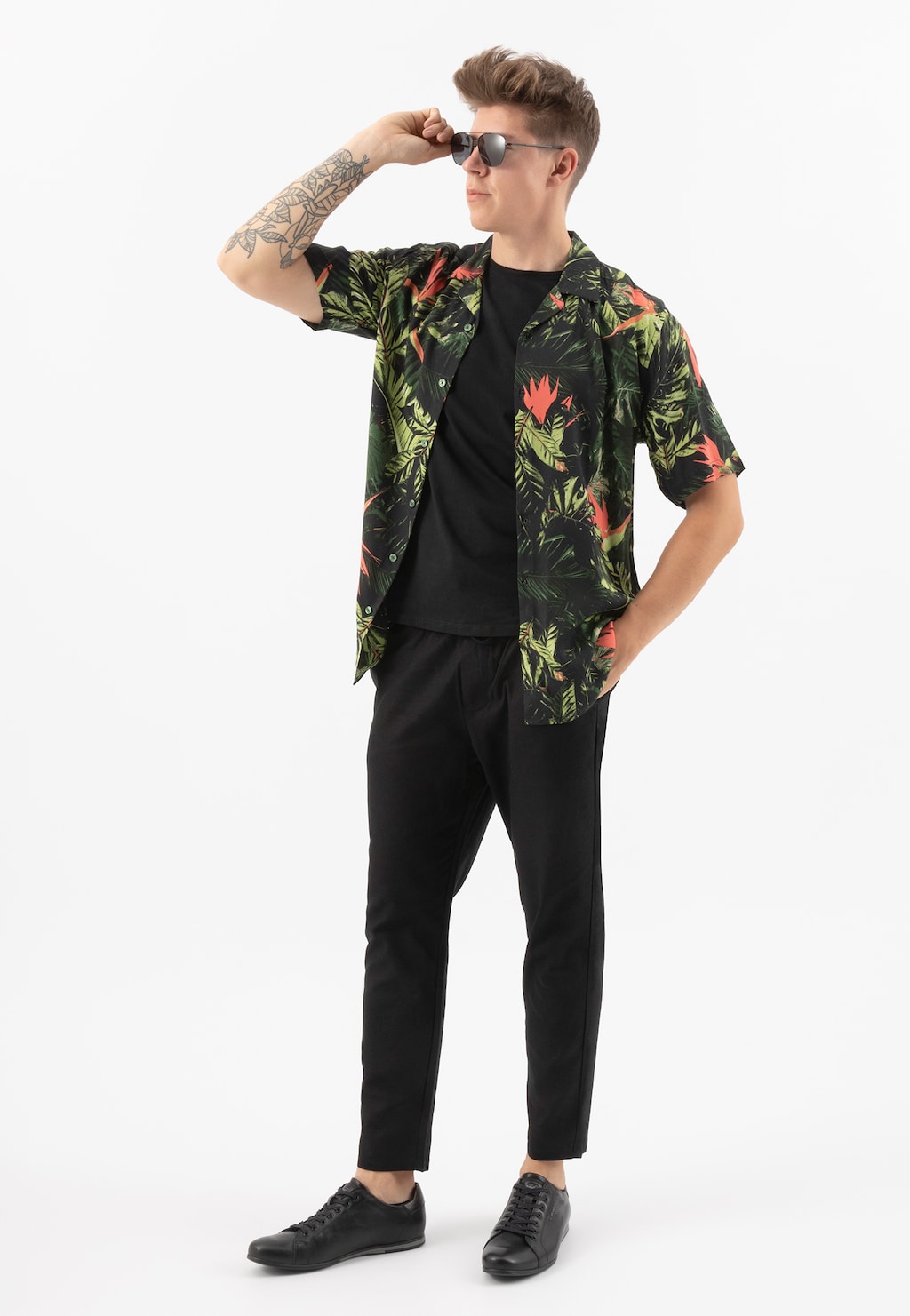 Mężczyzna w czarnych minimalistycznych sneakersach Wojas 8071-51, ubrany w kwiecistą koszulę i czarne spodnie, patrzący w dal z ręką przy okularach przeciwsłonecznych.