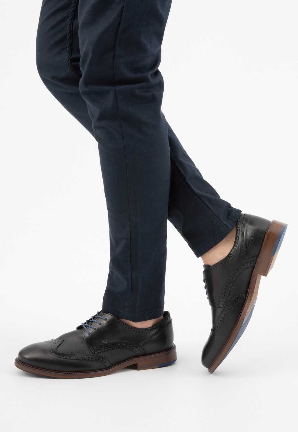 Eleganckie czarne półbuty męskie Wojas model 9072-51 typu brogue z perforacjami, brązową podeszwą i kontrastującym obcasem, prezentowane na nogach w granatowych spodniach.