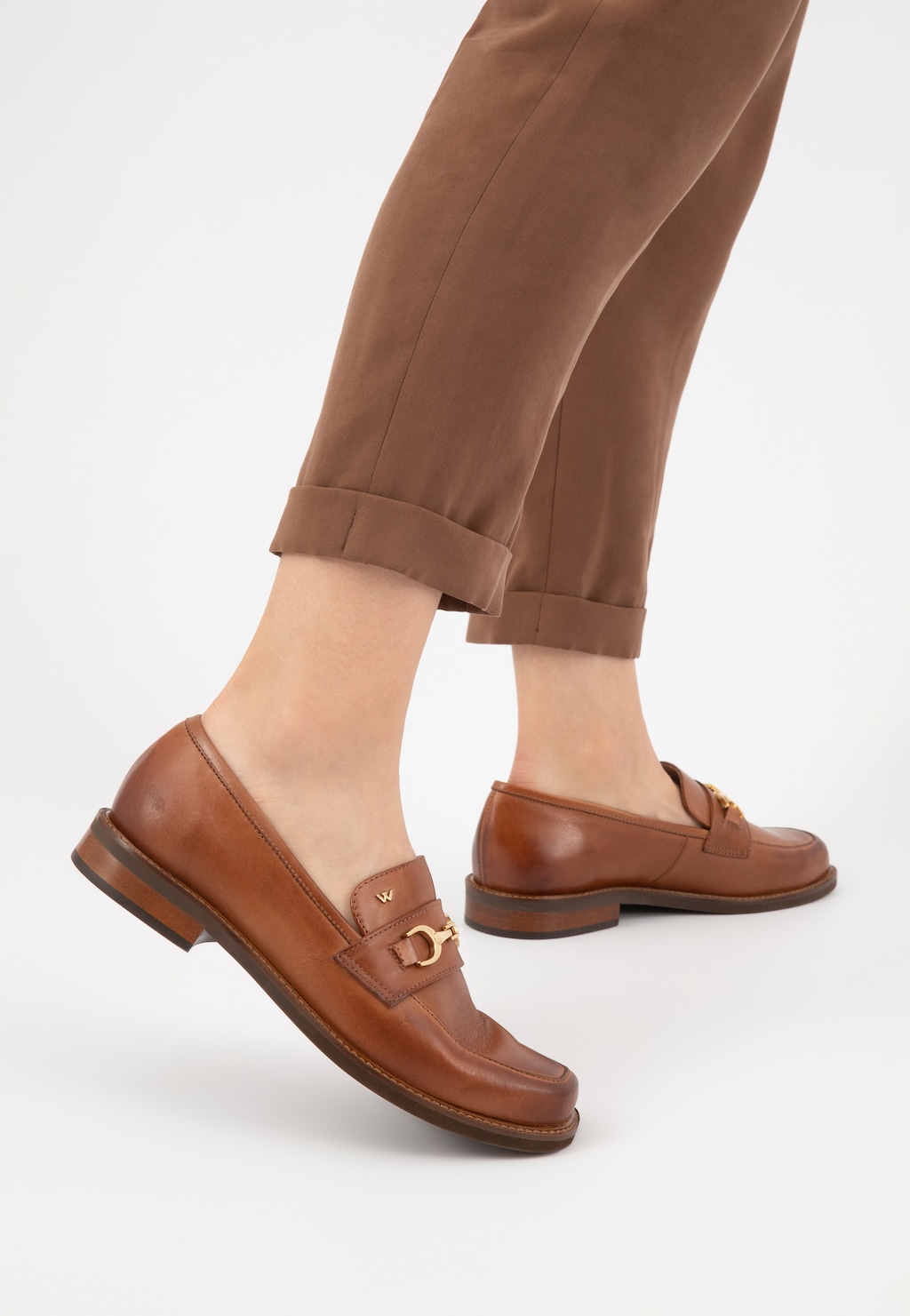 Eleganckie jasnobrązowe mokasyny Wojas model 46092-53 ze skóry licowej na stopach osoby w podwiniętych brązowych spodniach, widok z boku ukazujący złote zdobienie.