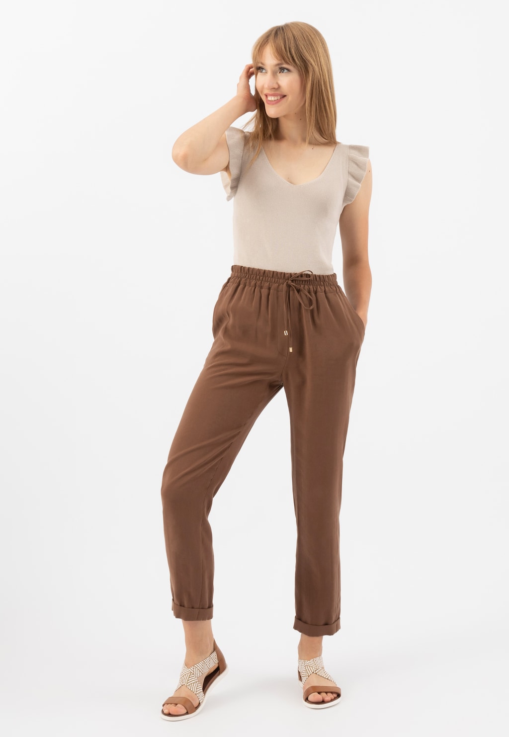 Uśmiechnięta kobieta w beżowej bluzce i brązowych spodniach, prezentująca jasnobrązowe sandały damskie Wojas z plecionymi paskami model 76018-83 na białym tle.