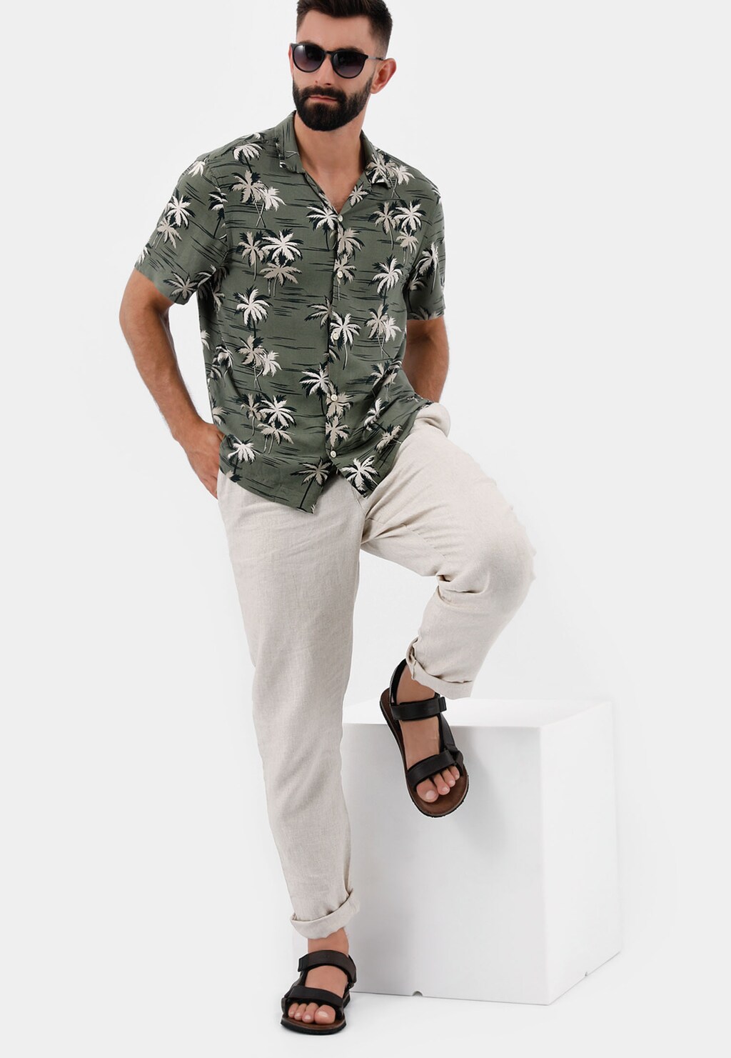 Osoba w zielonej koszuli w palmy, jasnych spodniach i brązowych sandałach męskich Wojas model 29007-52 z przeplatanymi paskami, pozująca z nogą na białym postumencie.