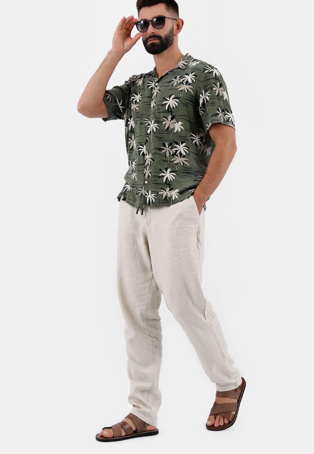 Mężczyzna w zielonej koszuli w palmy, jasnych spodniach i brązowych sandałach Wojas model 29007-52 z przeplatanymi paskami, dotykający okularów przeciwsłonecznych.