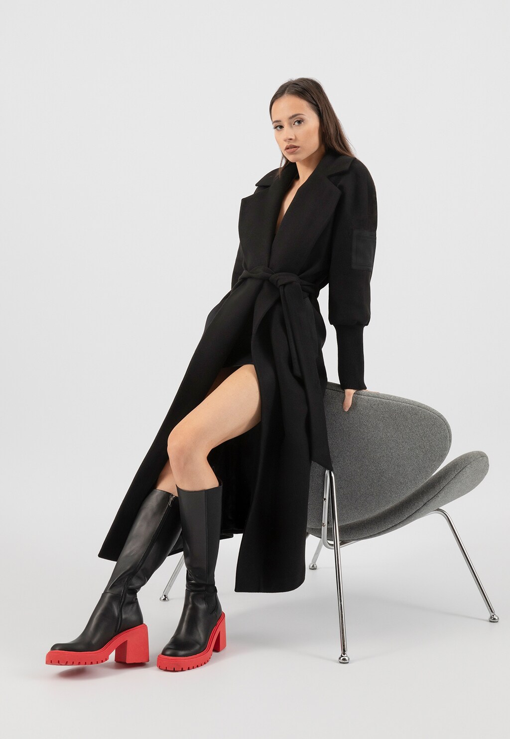 Czarne kozaki sztyblety Wojas 71110-51 z czerwoną podeszwą na obcasie, prezentowane na modelce w długim czarnym płaszczu, siedzącej na nowoczesnym szarym fotelu.