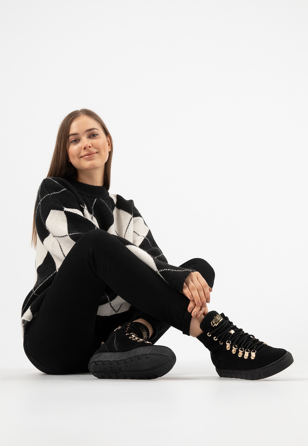 Kobieta w czarno-białym swetrze prezentująca czarne trzewiki damskie Relaks R64006-61 ze złotymi elementami, ocieplane buty widoczne na nodze w czarnych spodniach.