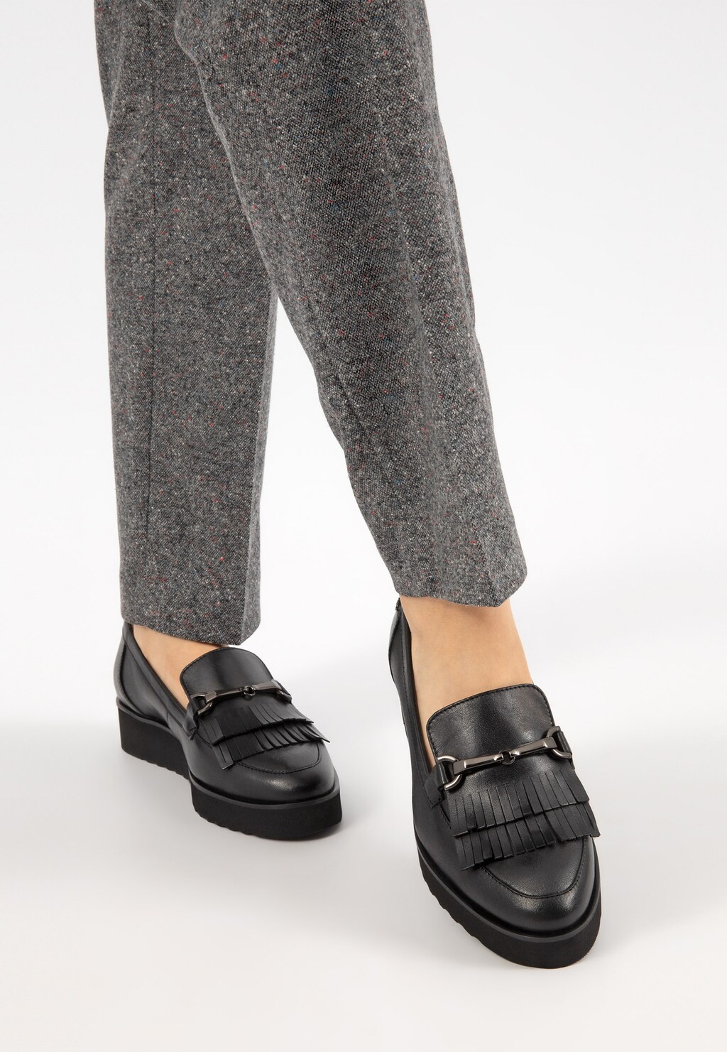 Czarne mokasyny damskie Wojas model 46100-51 z ozdobnymi frędzlami, noszone do szarych spodni. Widok z przodu na eleganckie buty z dekoracyjnymi elementami.