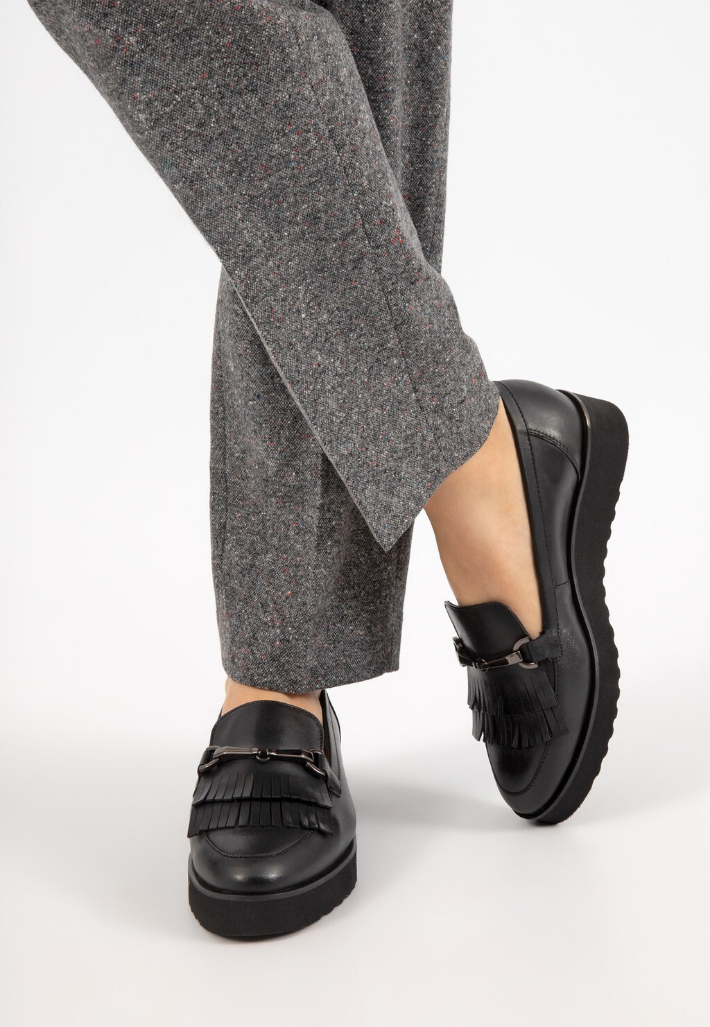 Czarne mokasyny damskie Wojas 46100-51 z ozdobnymi frędzlami na nodze osoby w szarych spodniach, widok z boku pokazujący elegancki design i praktyczność obuwia.