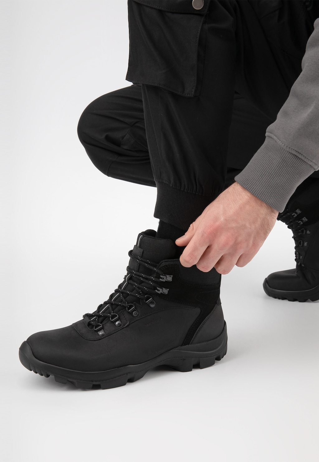 Czarne wysokie trzewiki trekkingowe Wojas model 24029-71 prezentowane na nodze osoby ubranej w czarne spodnie. Widok z boku pokazujący sznurowanie i masywną podeszwę buta.