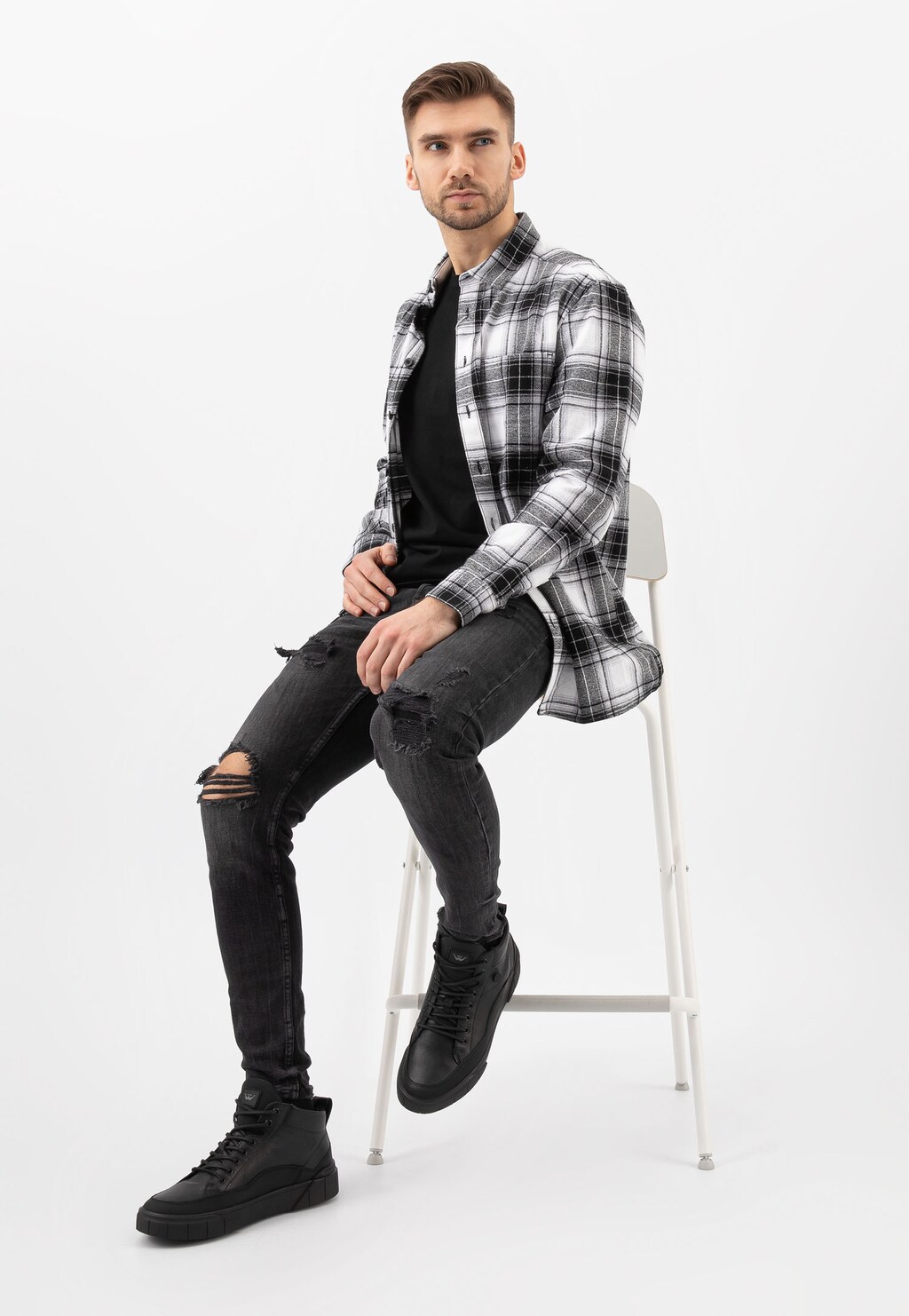 Mężczyzna w kraciastej koszuli siedzi na białym stołku, prezentując czarne skórzane trzewiki Wojas model 24067-71 - stylowe męskie trampki z ociepleniem.