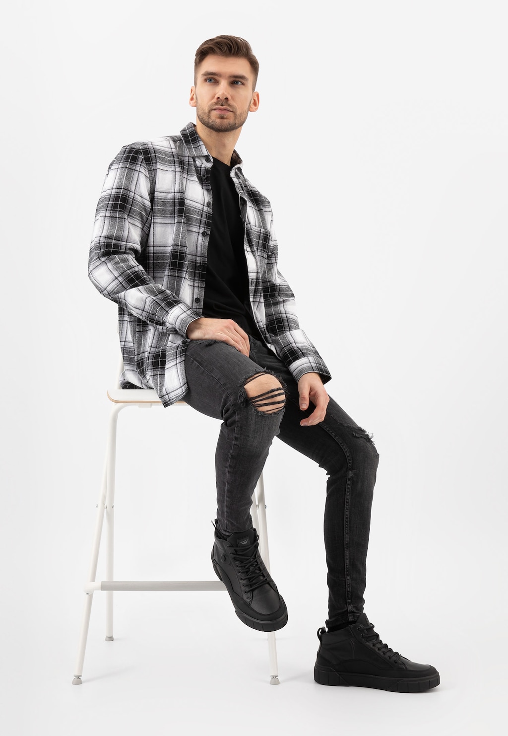 Mężczyzna w kraciastej koszuli i czarnym t-shircie, siedzący na białym stołku, ubrany w czarne trzewiki męskie Wojas model 24067-71, stylizowane na trampki z ociepleniem.