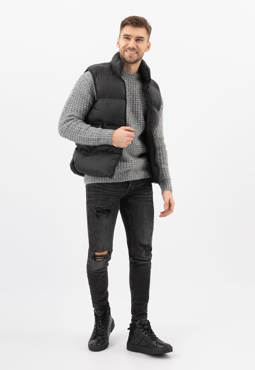 Mężczyzna w czarnym bezrękawniku i szarym swetrze, ubrany w ciemne jeansy i czarne trampki Wojas model 24070-71, uśmiecha się prezentując casualowy zimowy styl.