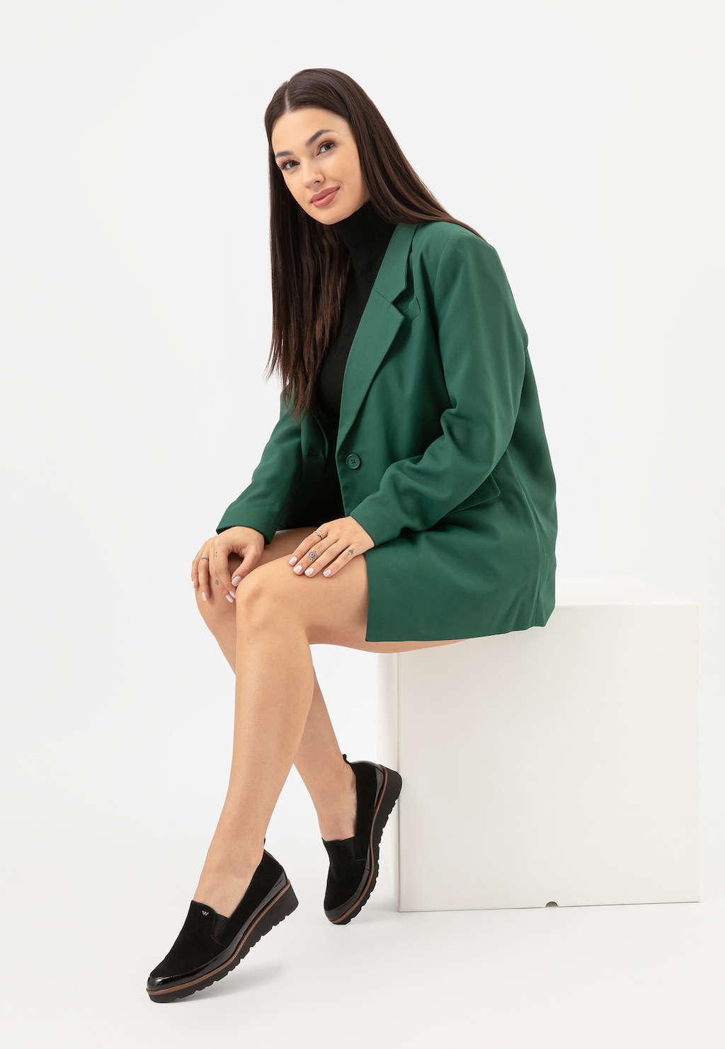 Elegancka modelka w zielonym płaszczu prezentuje czarne wsuwane półbuty damskie Wojas model 46008-71 z łączonych skór, siedząc na białym podium ze skrzyżowanymi nogami.