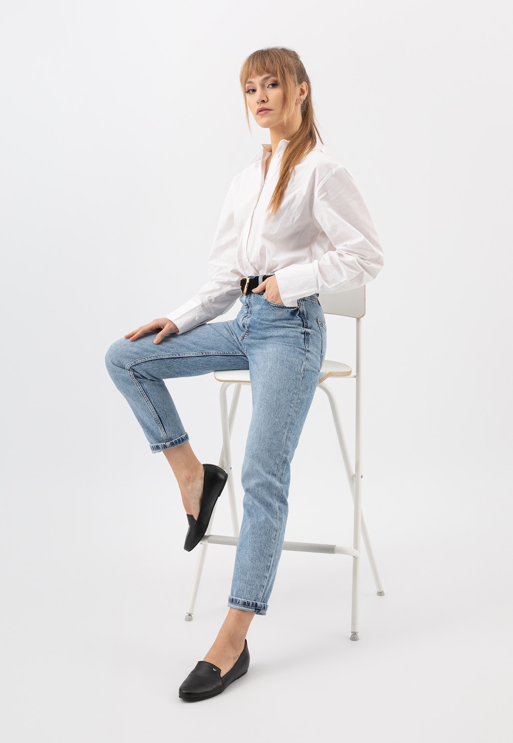 Modelka w białej koszuli i jeansach siedząca na krześle, prezentująca czarne lordsy damskie Wojas model 45003-50 na płaskiej podeszwie, ukazane z bocznej perspektywy.