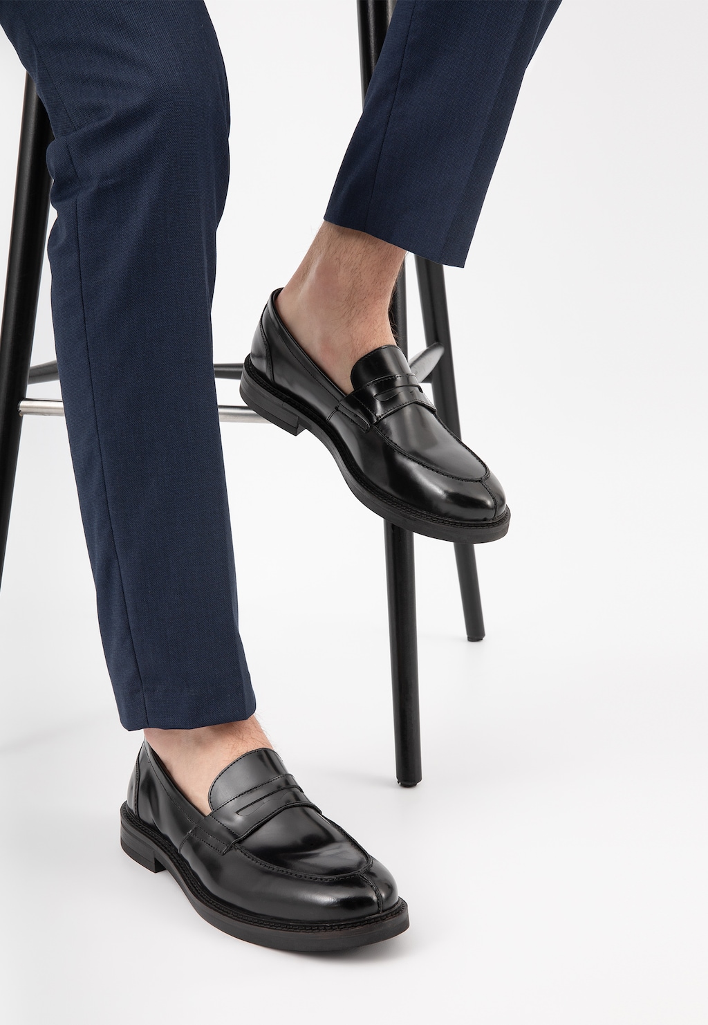 Czarne lakierowane mokasyny męskie Wojas 10137-31 typu penny loafers noszone z granatowymi spodniami, pokazane na modelu siedzącym na wysokim stołku.