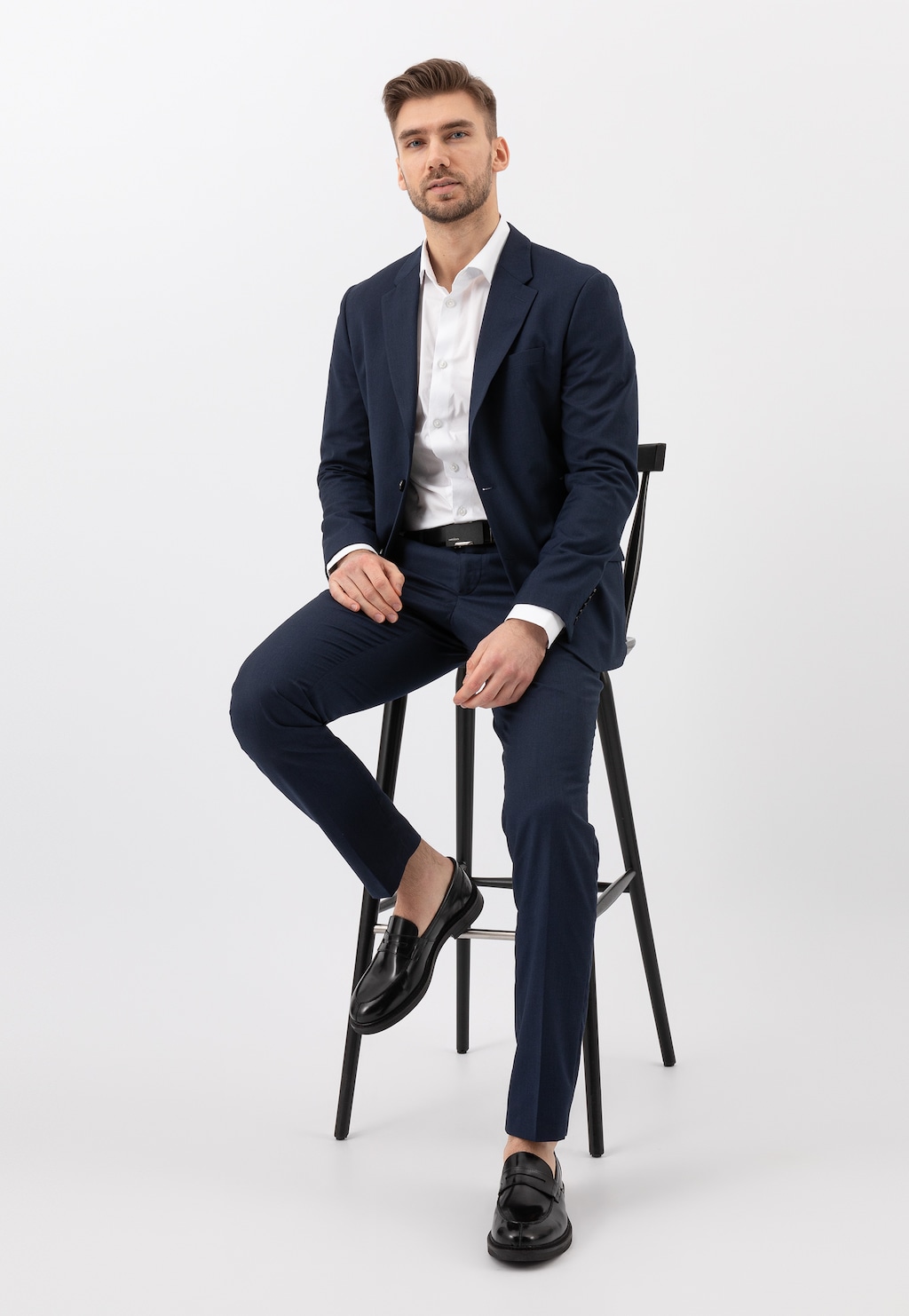 Elegancki mężczyzna w granatowym garniturze siedzący na krześle, prezentujący czarne lakierowane mokasyny męskie Wojas model 10137-31 typu penny loafers.