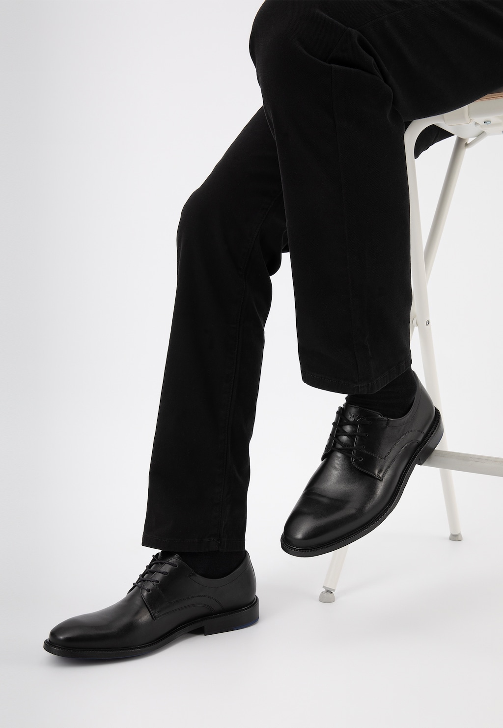 Eleganckie czarne półbuty męskie Wojas model 10133-51 na nogach osoby w czarnych spodniach, siedzącej na białym krześle. Klasyczne sznurowane obuwie w biznesowym stylu.