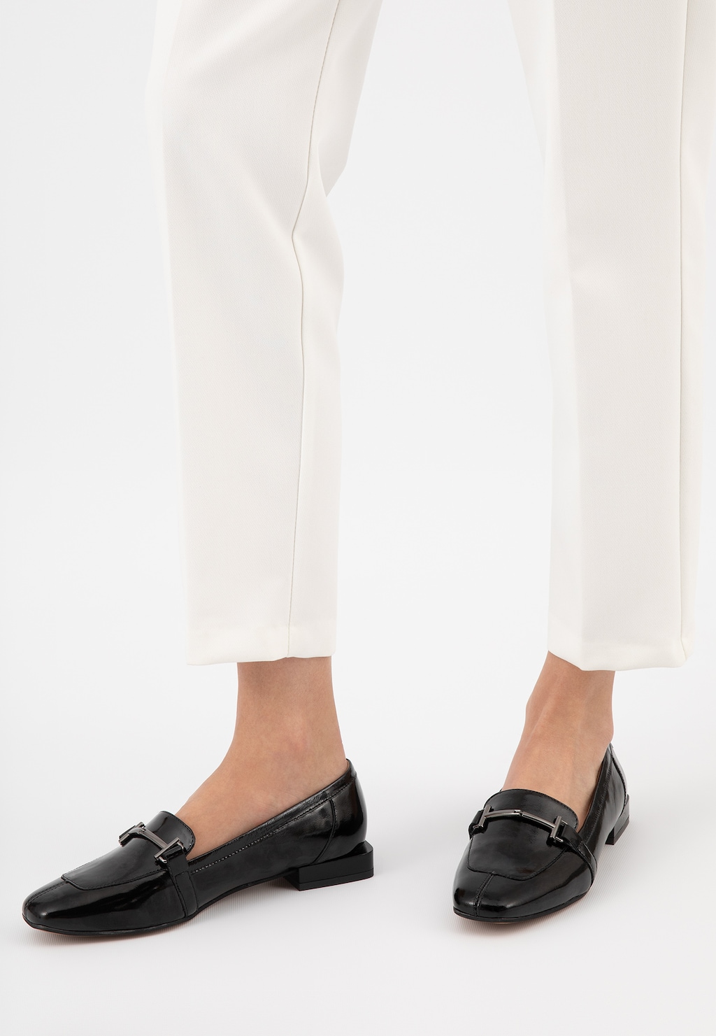 Czarne lakierowane mokasyny damskie Wojas 46223-31 z metalową ozdobą na przodzie, prezentowane na stopach modelki w białych spodniach, widok z boku.