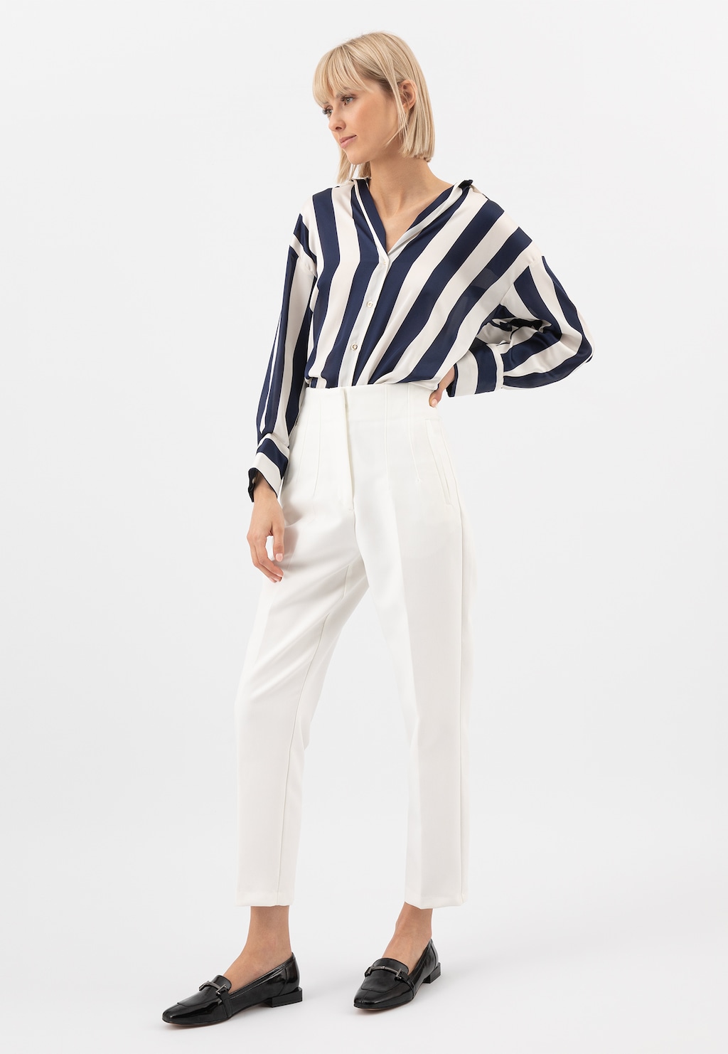 Kobieta w eleganckim stroju z czarno-białą pasiastą bluzką i białymi spodniami, prezentująca czarne lakierowane mokasyny damskie Wojas model 46223-31 z metalową ozdobą.