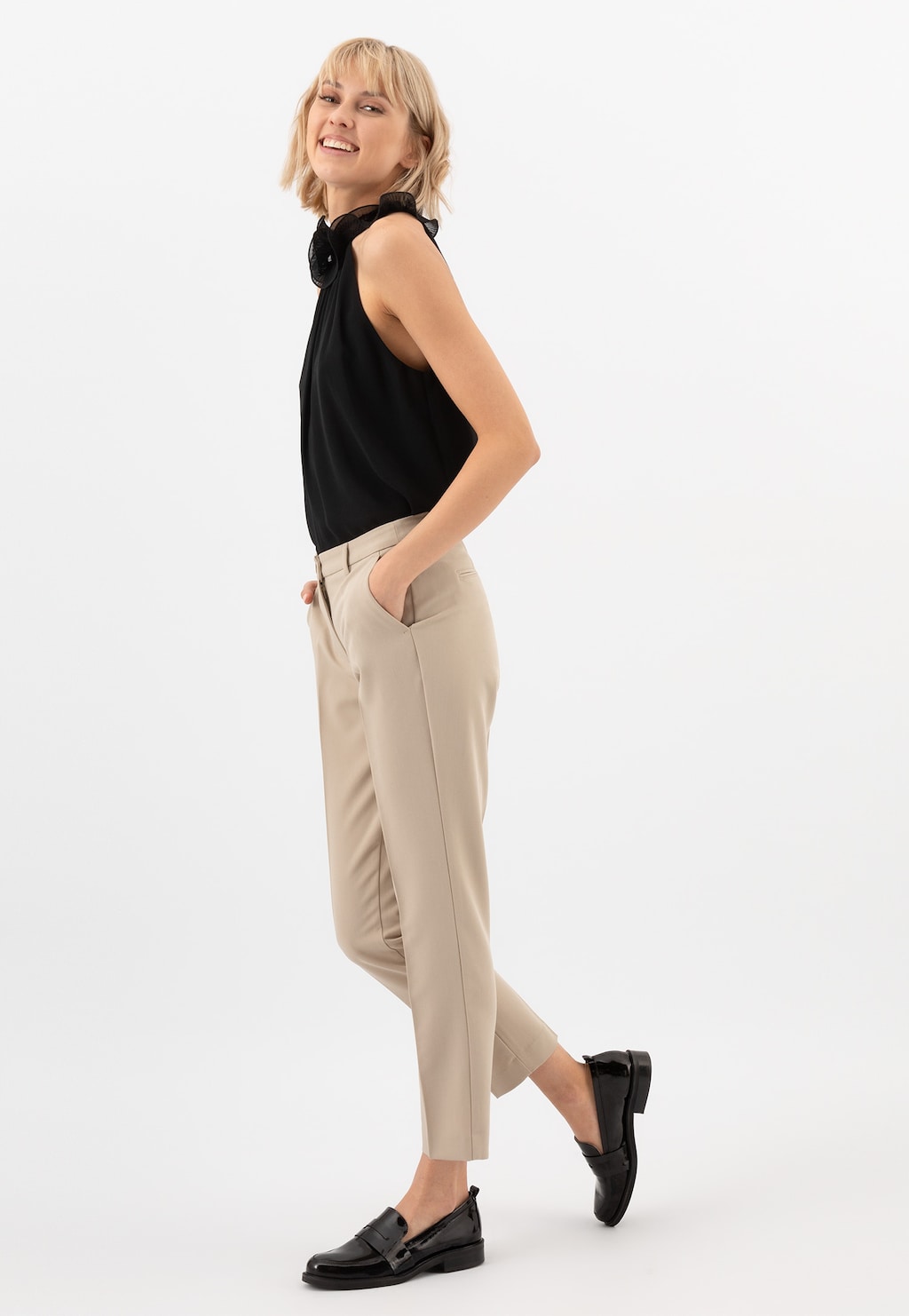 Kobieta w czarnej bluzce i beżowych spodniach prezentuje czarne lakierowane mokasyny damskie Wojas 46069-31 w stylu formal, widok z boku podkreśla elegancki profil obuwia.