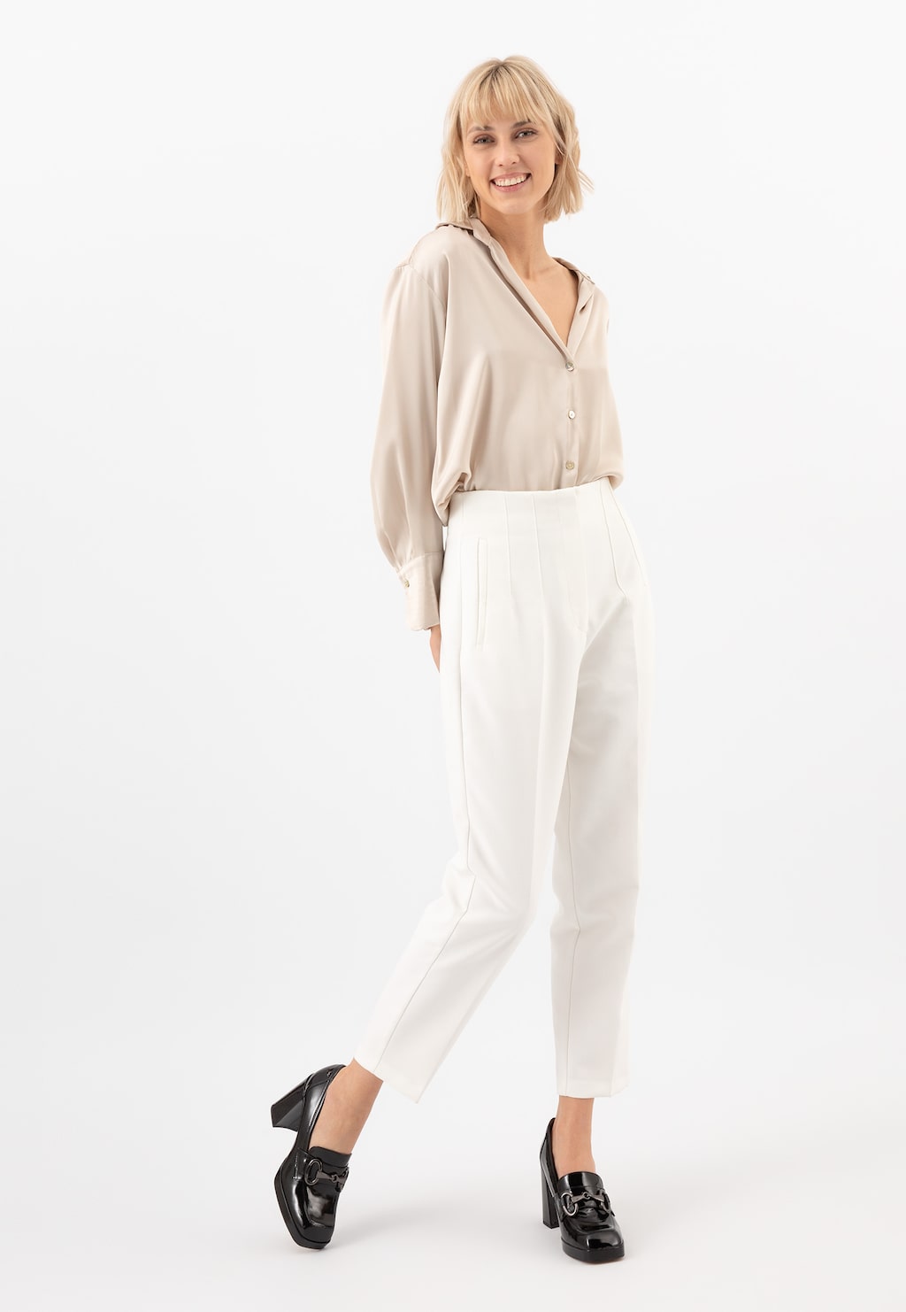 Uśmiechnięta kobieta w beżowej koszuli i białych spodniach prezentująca wysokie lakierowane mokasyny damskie Wojas na słupku model 46216-31, w eleganckim casualowym stylu.