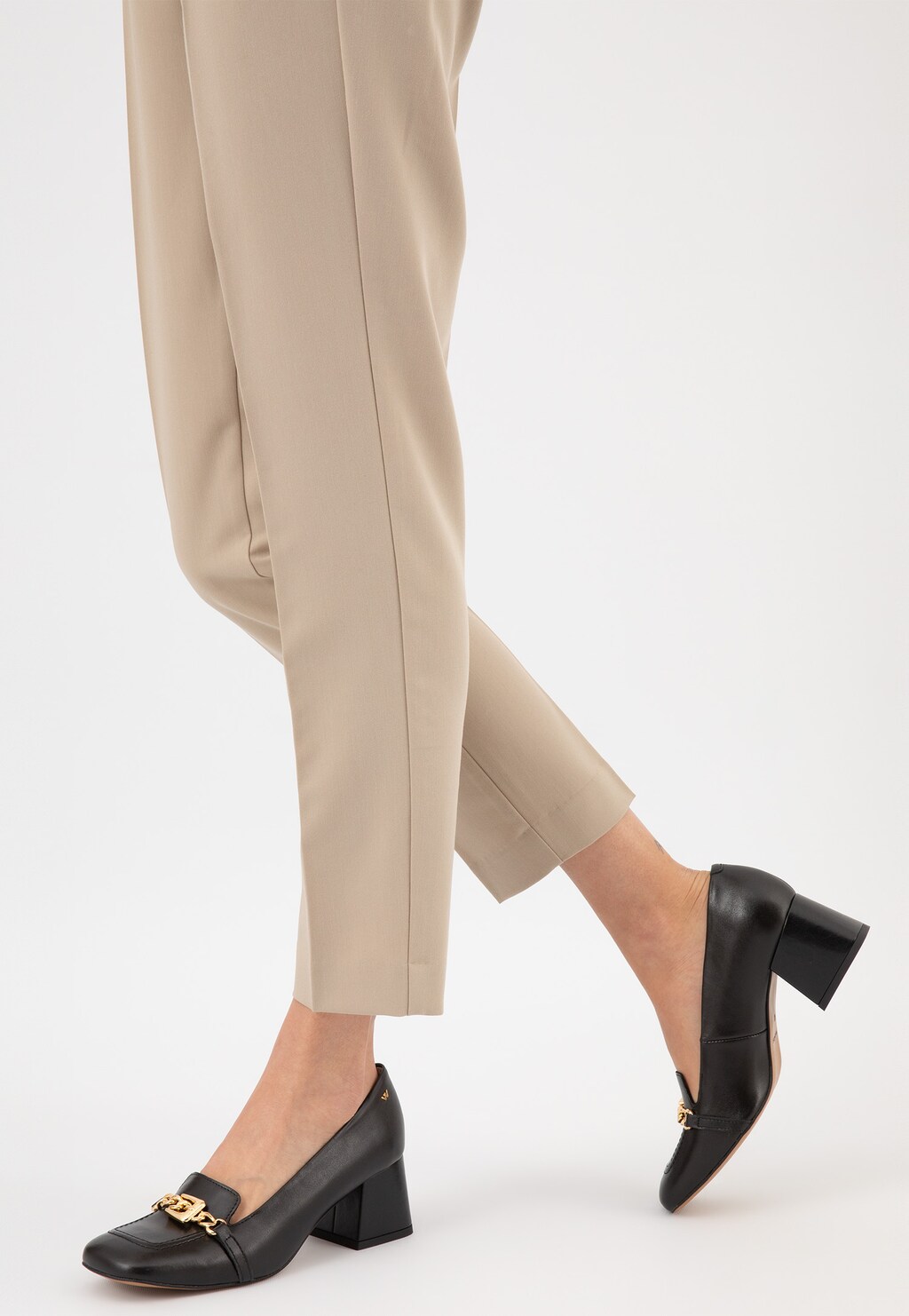Czarne skórzane mokasyny damskie Wojas 46237-51 na stabilnym obcasie ze złotą metalową ozdobą, prezentowane na nodze modelki w beżowych spodniach.