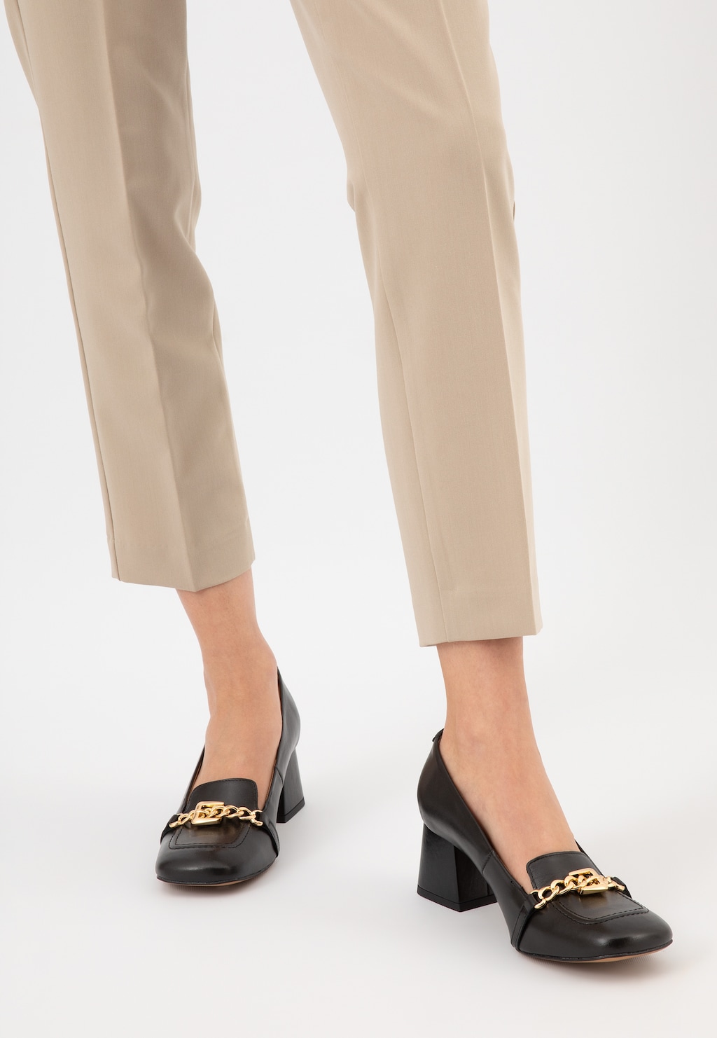 Czarne skórzane mokasyny damskie Wojas 46237-51 na stabilnym obcasie ze złotą ozdobą, prezentowane na nogach modelki ubranej w beżowe eleganckie spodnie.