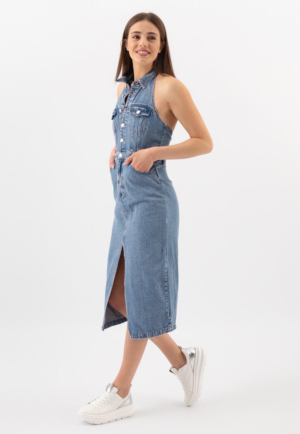 Modelka w jeansowej sukience bez rękawów prezentuje białe sneakersy Wojas 46228-59 ze srebrnymi wstawkami. Stylowe buty uzupełniają casualowy dżinsowy look.