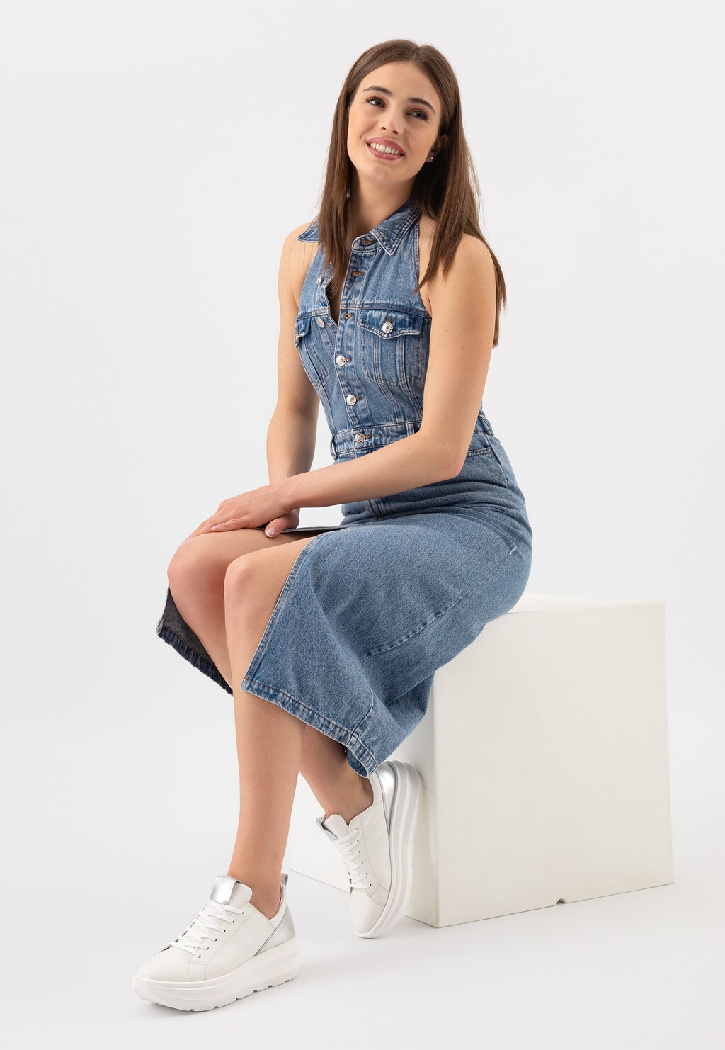 Modelka w jeansowej sukience prezentuje białe sneakersy Wojas 46228-59 ze srebrnymi wstawkami. Obuwie widoczne z profilu na białej podstawie.