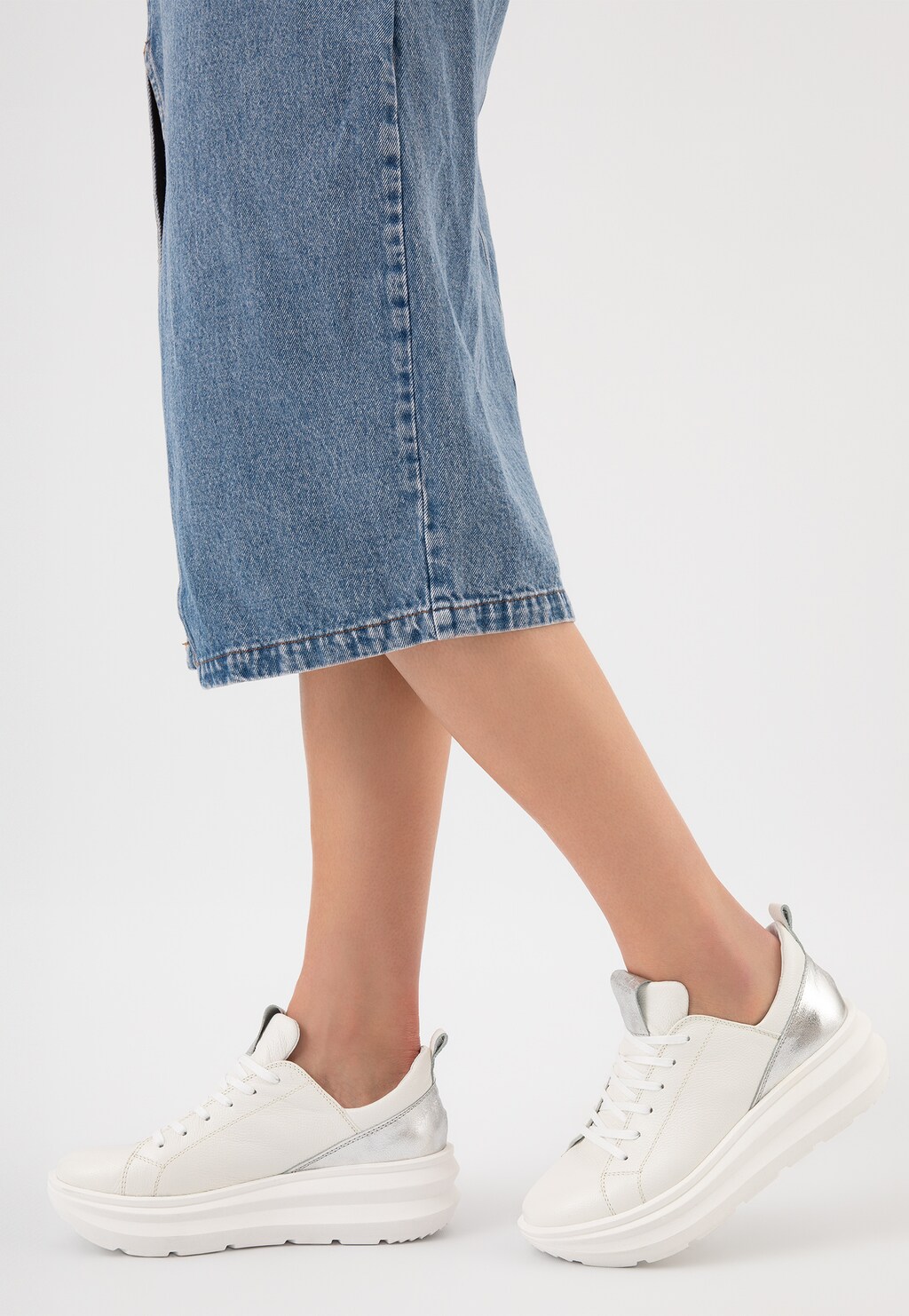 Białe sneakersy damskie Wojas 46228-59 ze srebrnymi wstawkami na nogach modelki w jeansowej spódnicy. Buty prezentują się z boku, mają grubą białą podeszwę.