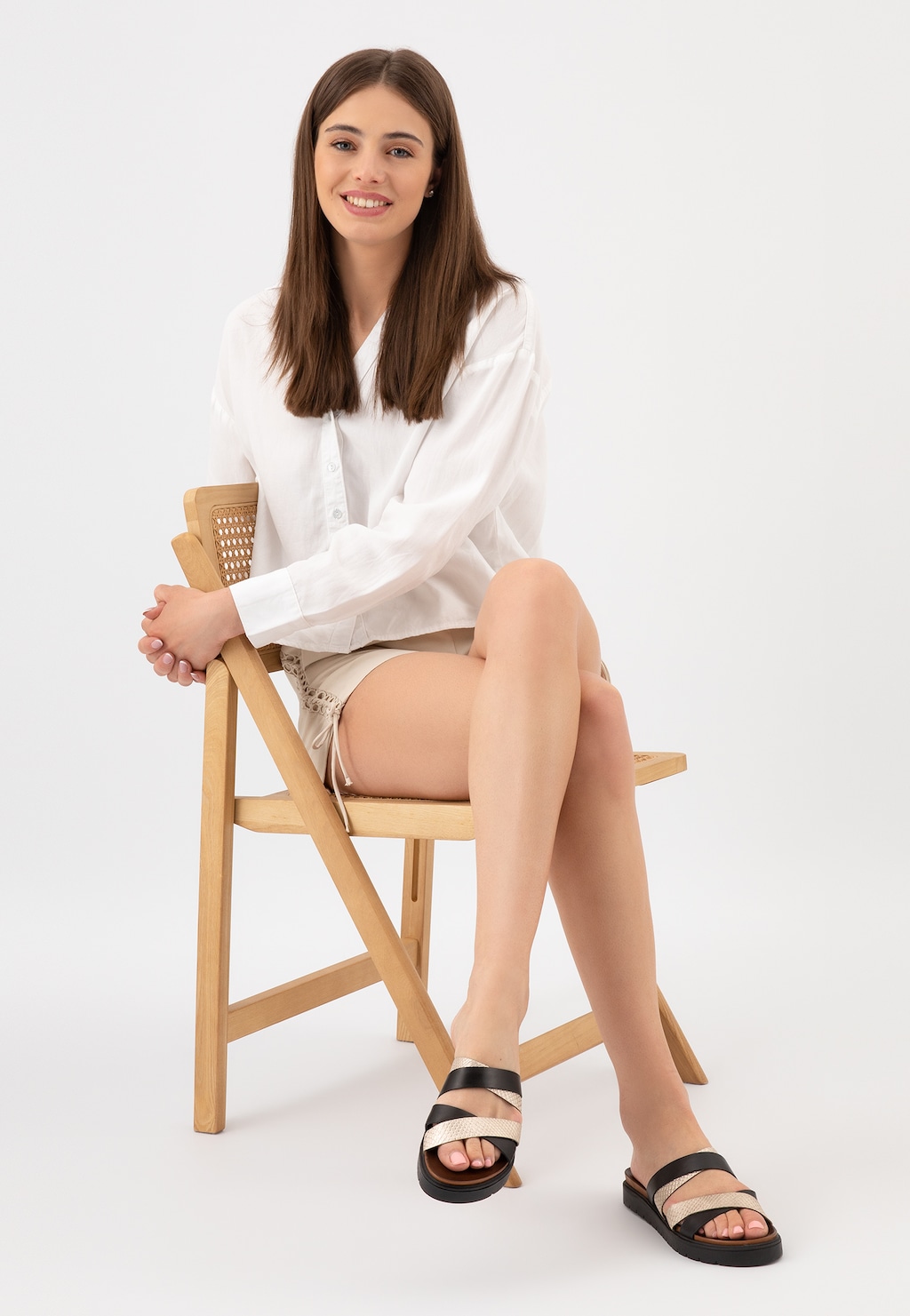 Modelka w białej koszuli i jasnych szortach prezentuje czarne klapki damskie Wojas 74109-71 ze złotymi paskami, siedząc na drewnianym krześle.