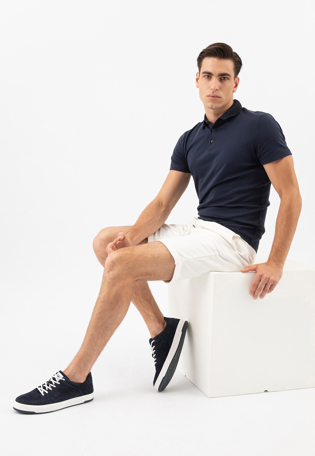 Model w granatowej koszulce polo i białych szortach siedzi na kostce, prezentując granatowo-białe półbuty męskie Wojas model 10151-66 typu trampki z białymi sznurówkami.
