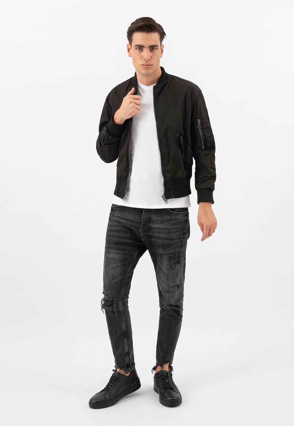 Model w modnej stylizacji prezentuje czarne trampki Wojas 10149-51 ze skóry licowej z zamkiem na cholewce, zestawione z ciemnymi jeansami, białym t-shirtem i czarną kurtką bomberką.