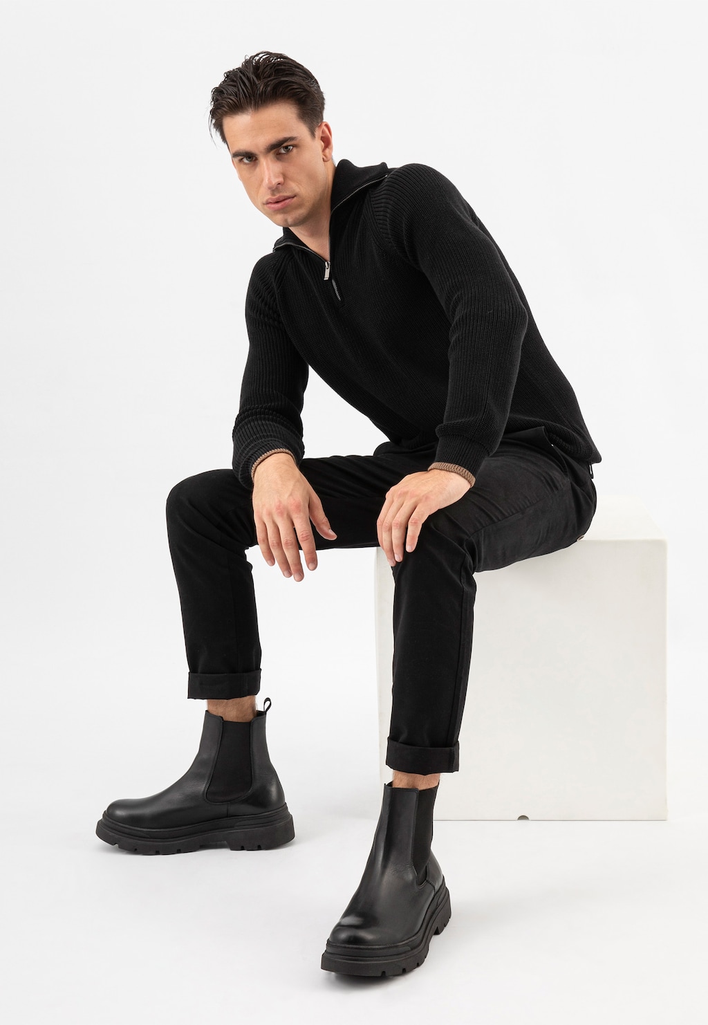 Model w czarnym stroju prezentujący czarne sztyblety męskie Wojas 20013-51 na grubej podeszwie, siedzący na białym postumencie w eleganckiej stylizacji.