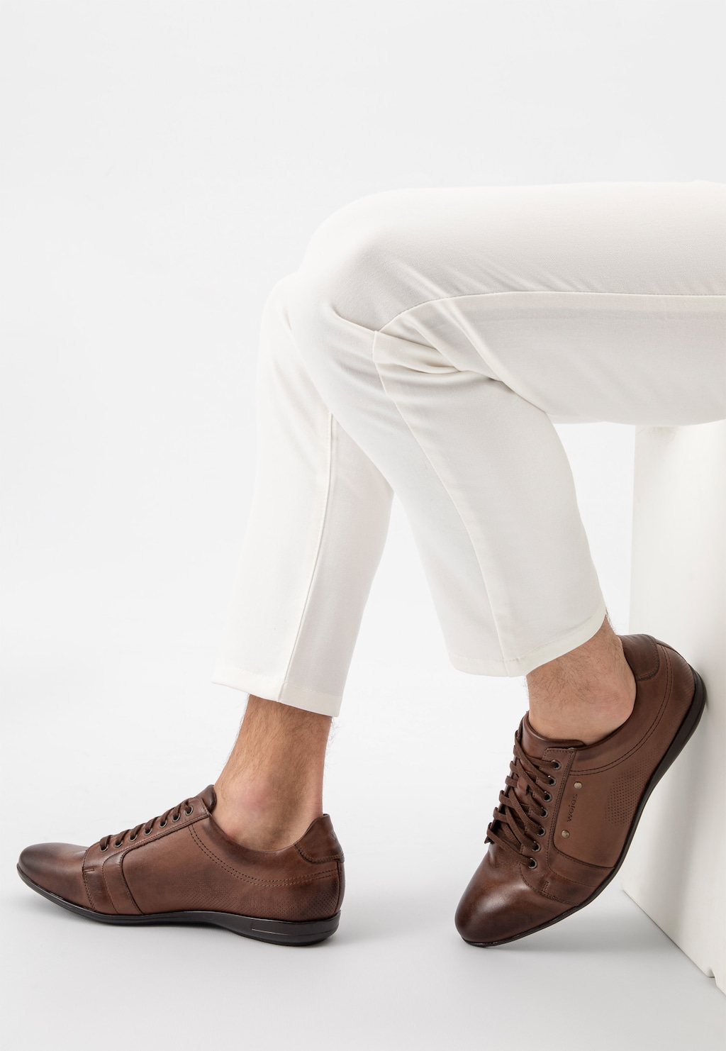 Eleganckie brązowe półbuty męskie Wojas model 7004-52 noszone z białymi spodniami, ukazane z bocznej perspektywy na nodze mężczyzny siedzącego na białej sofie.
