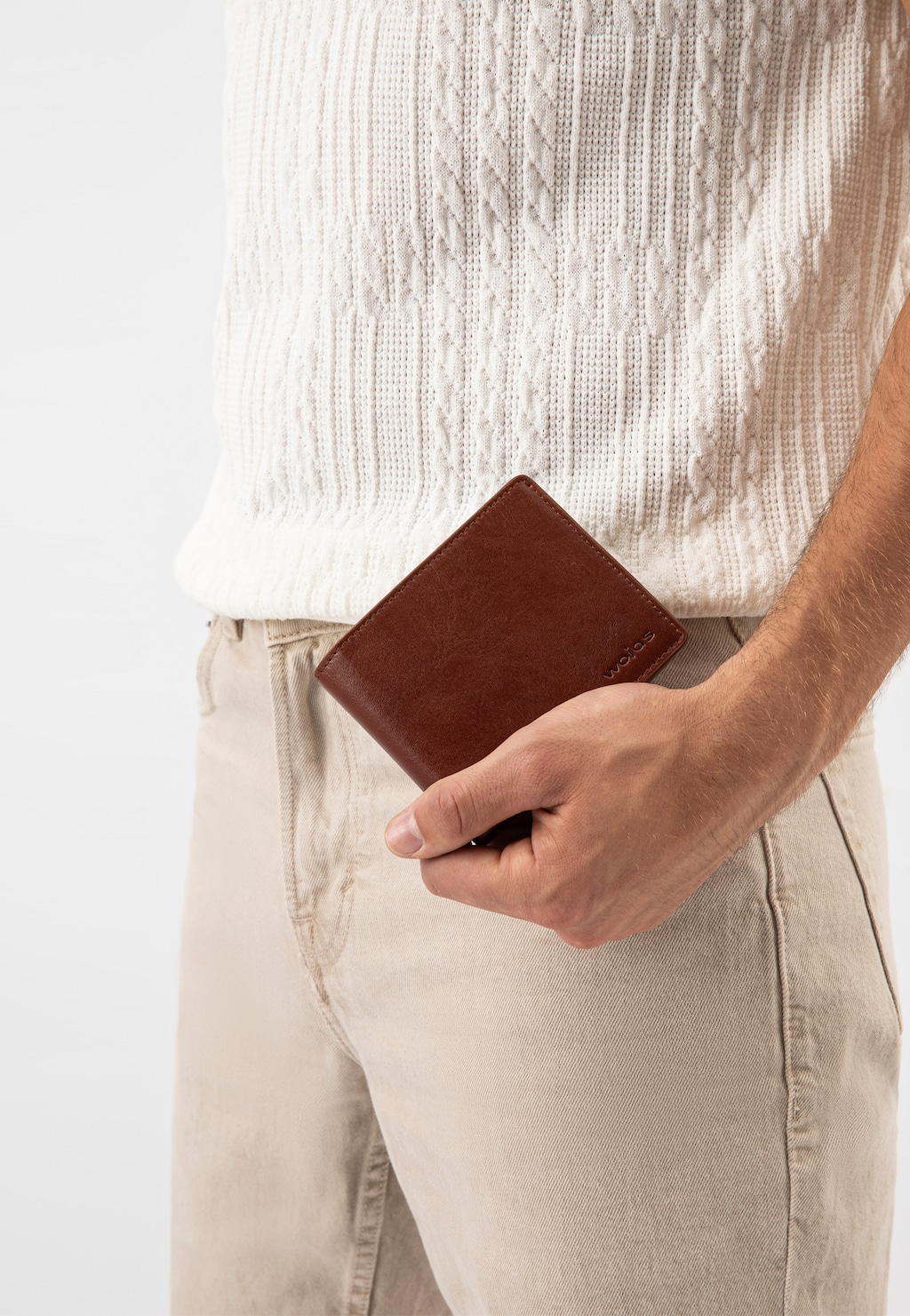 Dłoń mężczyzny trzymająca jasnobrązowy skórzany portfel Wojas model 91072-53. Elegancki produkt uchwycony przy biodrze, na tle białego swetra i beżowych spodni.
