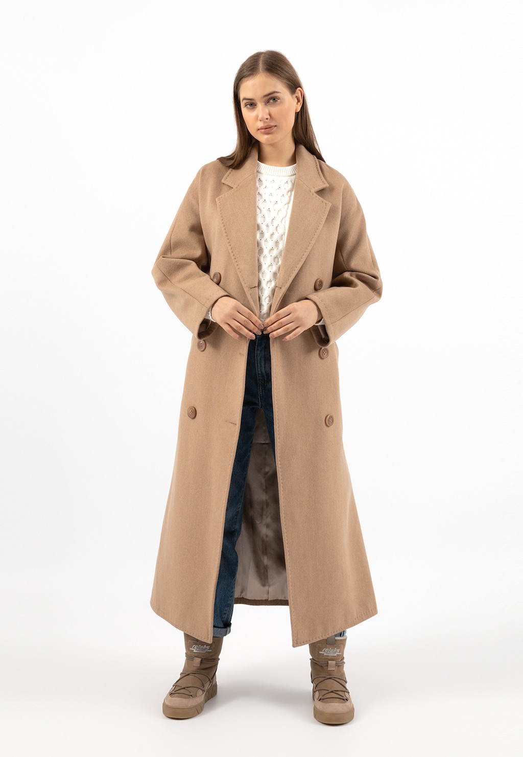 Elegancki beżowy płaszcz damski Relaks R99015-44, pokazany na modelce w białym swetrze i jeansach. Długi krój z klasycznym kołnierzem i podwójnym rzędem guzików.