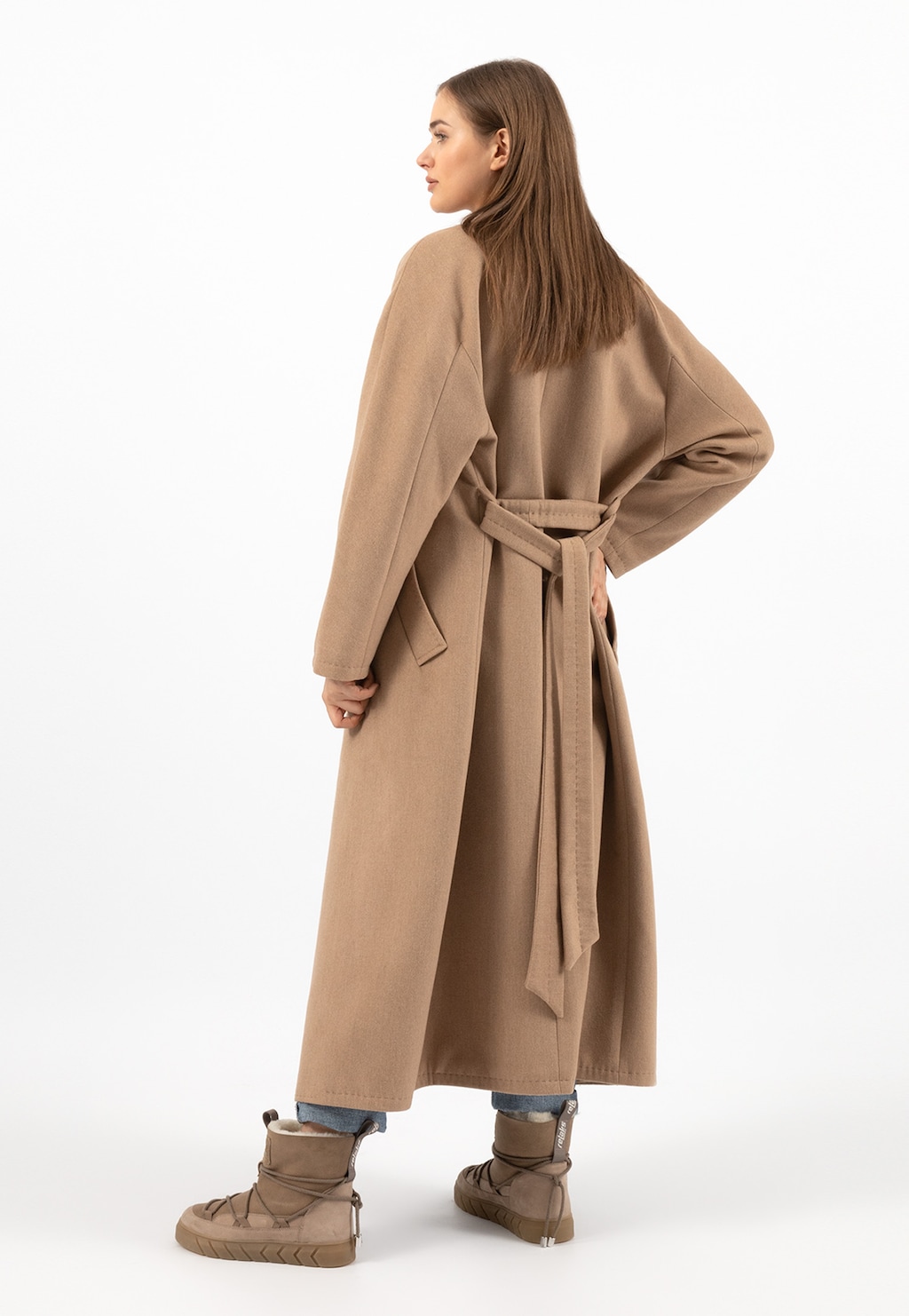Elegancki długi płaszcz damski Relaks R99015-44 w kolorze beżowym, widok z tyłu, ukazujący pasek w talii, noszonego z jeansami i brązowymi butami.