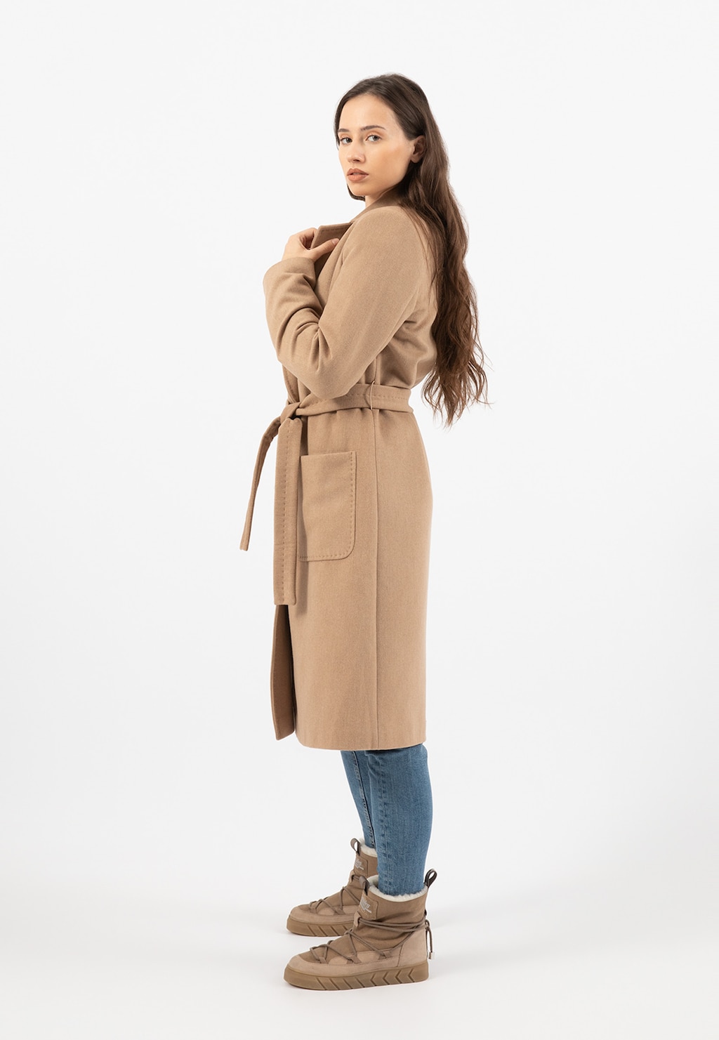 Elegancki beżowy płaszcz damski RELAKS model R99020-44 z paskiem, kieszeniami i rozcięciem, prezentowany w ujęciu z boku, noszony z jeansami i beżowymi butami.
