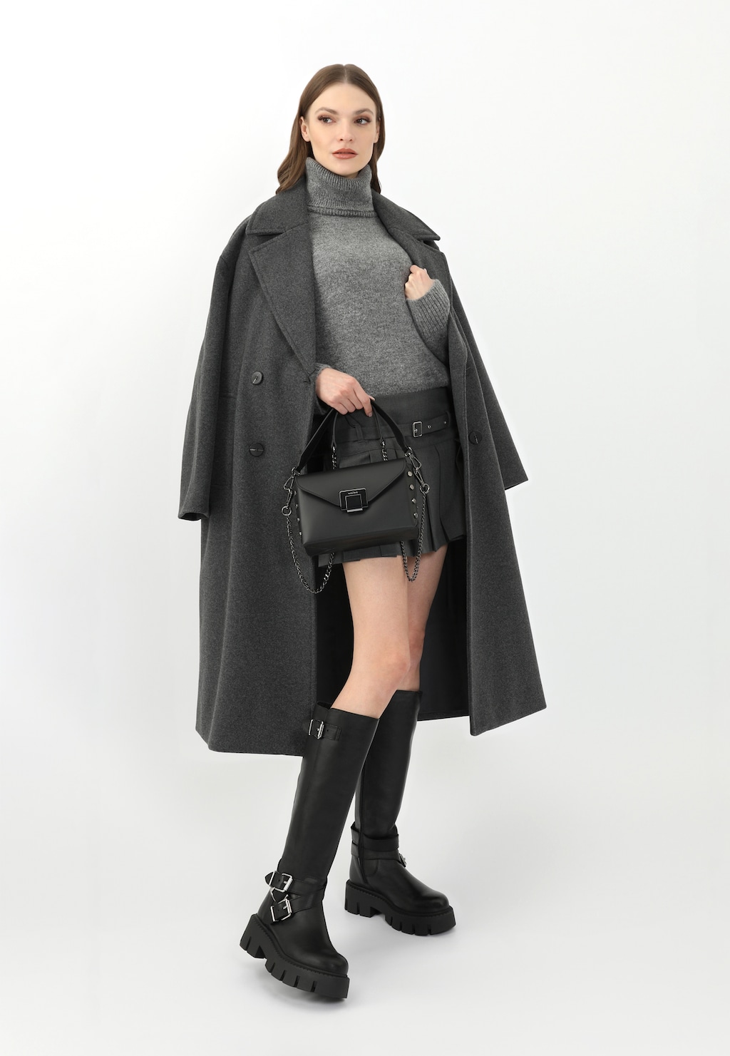 Modelka w szarym płaszczu i swetrze prezentująca czarne skórzane kozaki damskie Wojas typu bikery ze srebrnymi klamrami model 71053-51, z czarną torebką.