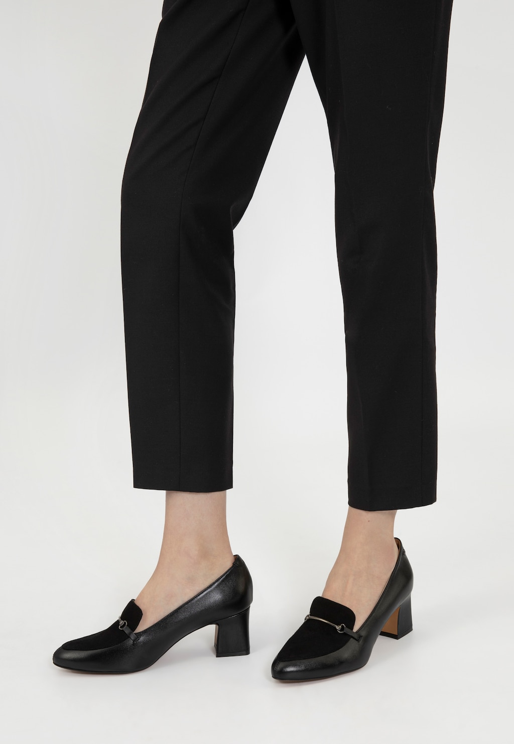 Eleganckie czarne półbuty damskie Wojas model 46014-71 na stabilnym obcasie, noszone z czarnymi spodniami. Buty mają ozdobny metalowy element na przodzie.