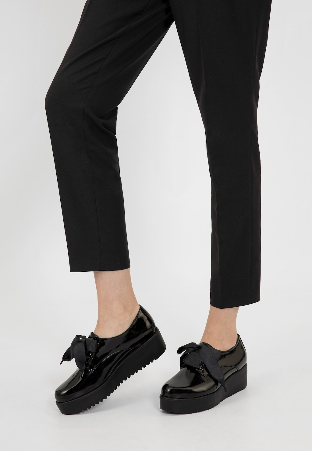 Czarne lakierowane półbuty damskie Wojas 46043-31 na platformie z wiązaniem, noszone z eleganckimi czarnymi spodniami. Widok z boku podkreśla modny charakter obuwia.