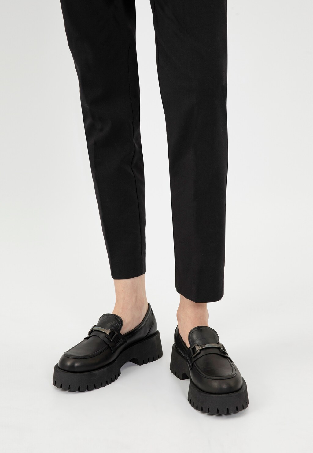 Czarne mokasyny damskie Wojas model 46198-51 z metalową ozdobą horsebit na nogach modelki w czarnych spodniach. Buty posiadają masywną, traktorową podeszwę.